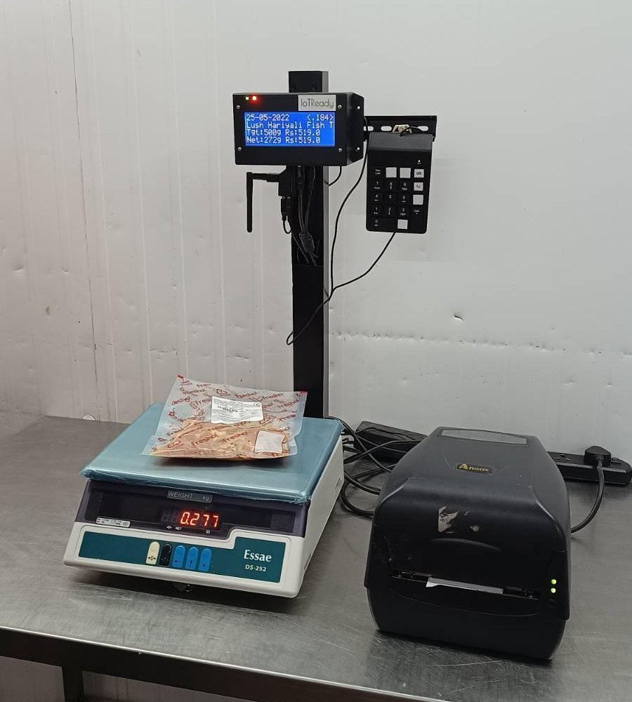 IoTReady label controller with a four-line display and keypad on a post above an Essae bench scale holding a packed bag, next to an Argox barcode printer